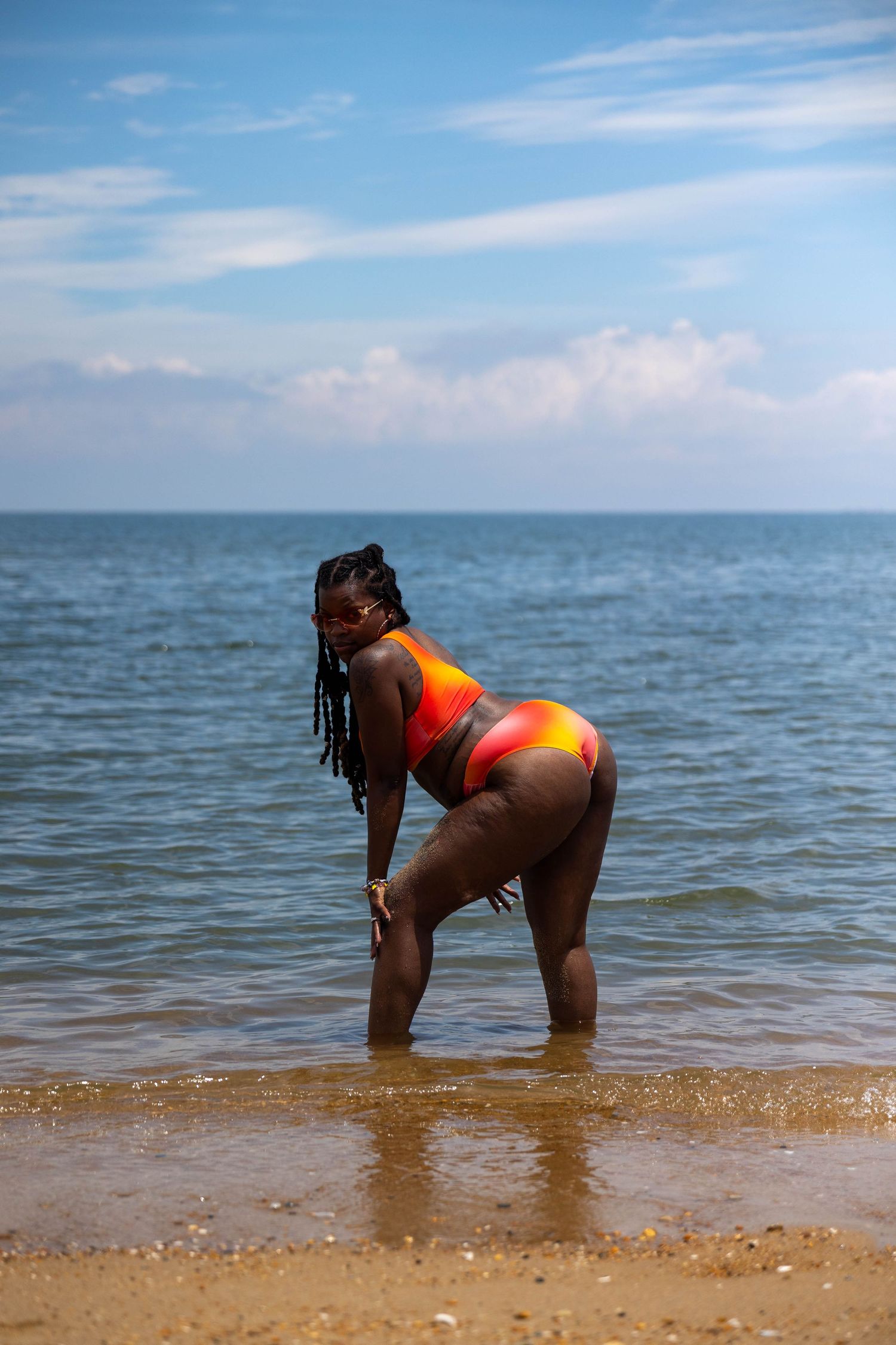 Woman in colorful orange bikini posing on sandy beach shoreline with calm blue ocean and cloudy sky backdrop.