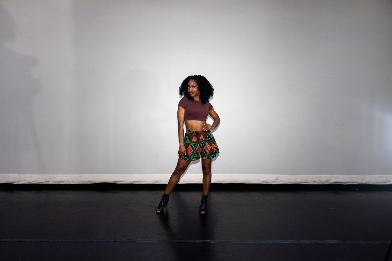 Young Black woman in colorful African print skirt and crop top poses on a dark stage floor.