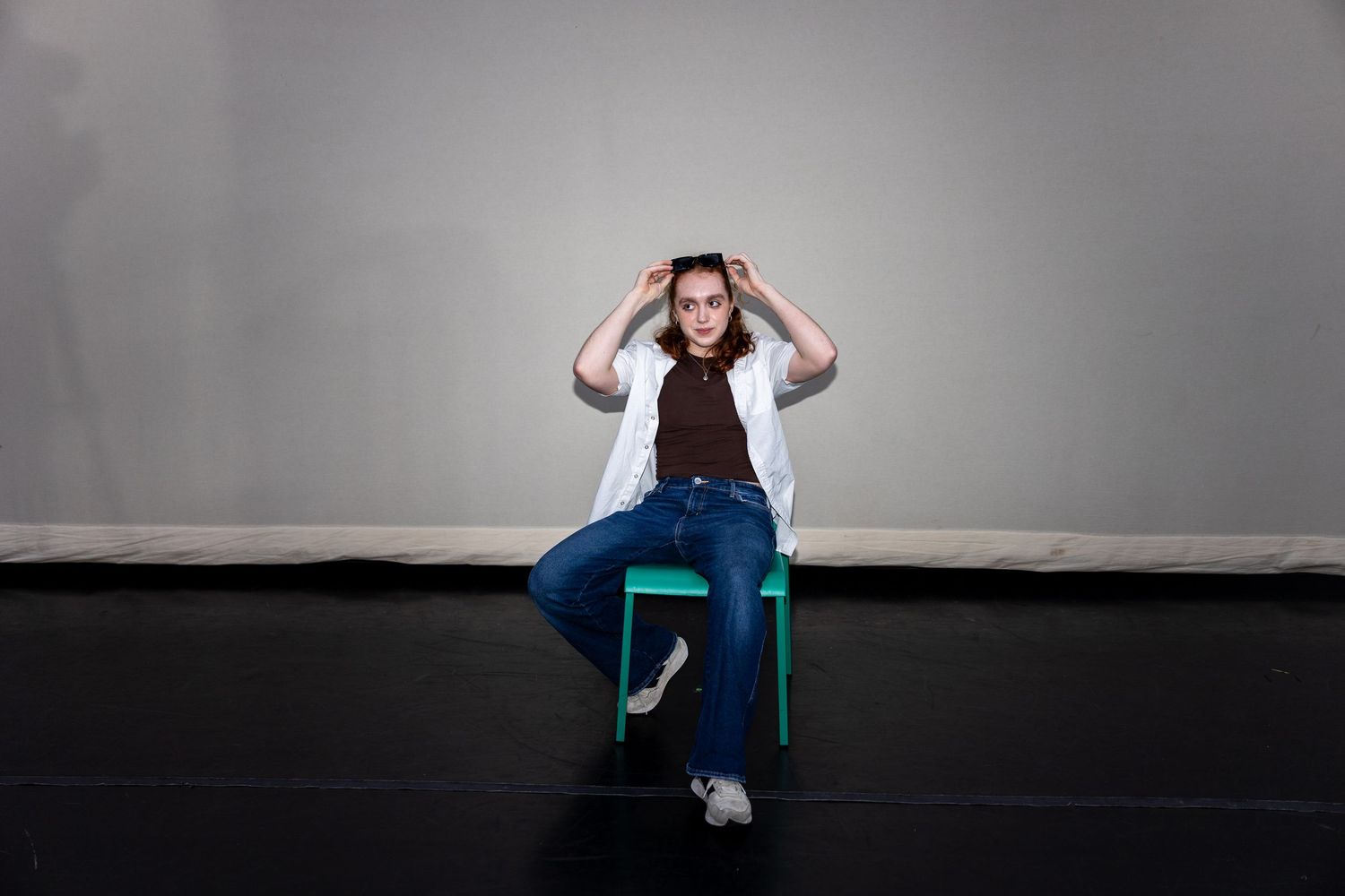 Woman in white blazer and jeans sitting on green chair with hands on head.
