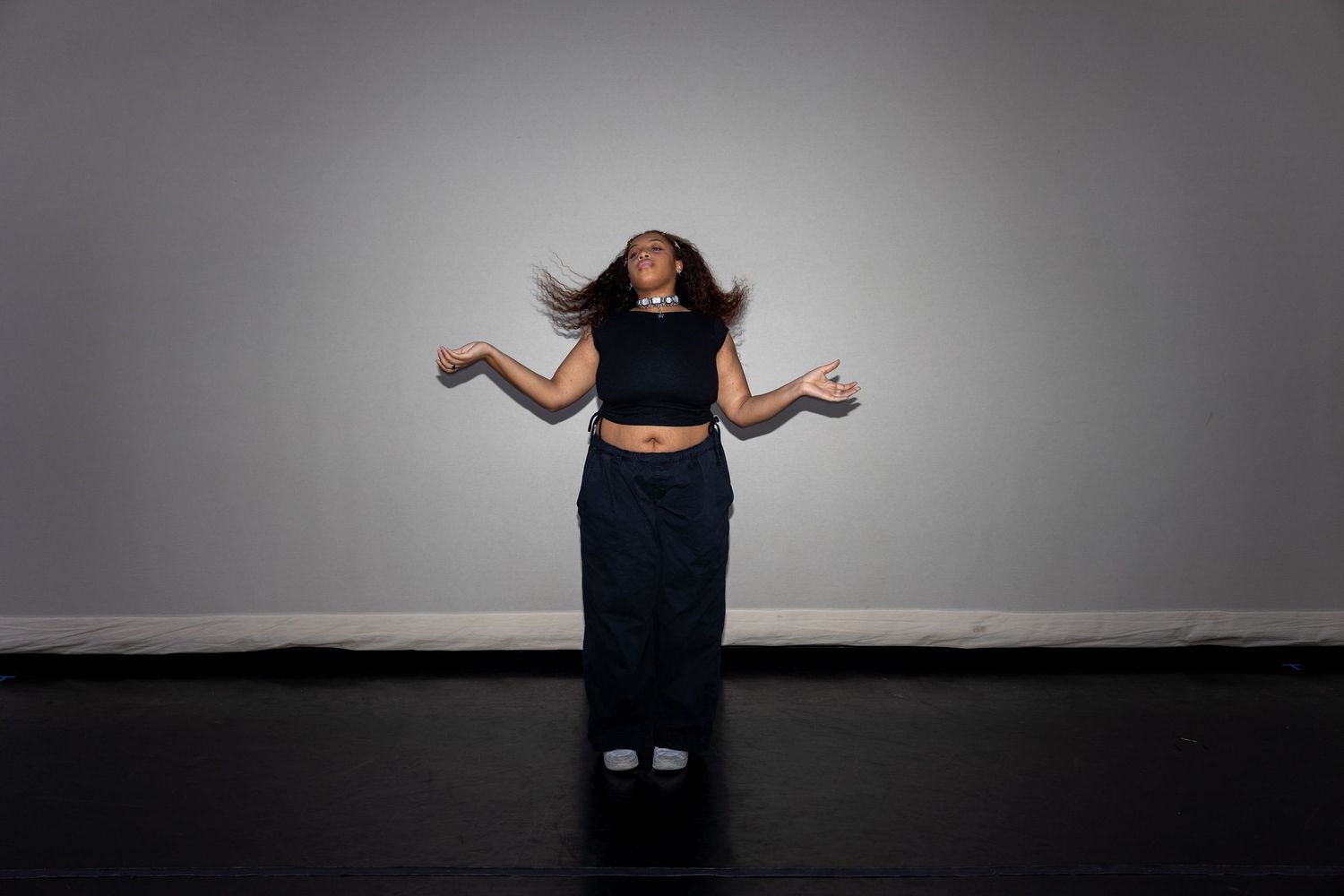 A dancer in a black crop top and navy skirt tosses her hair mid-movement in a gray studio setting.
