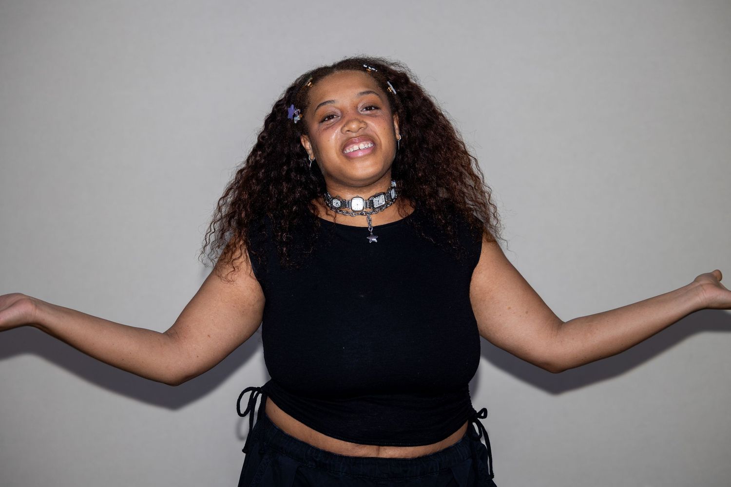 A young woman with curly hair in a black crop top and choker spreads her arms wide while laughing joyfully.