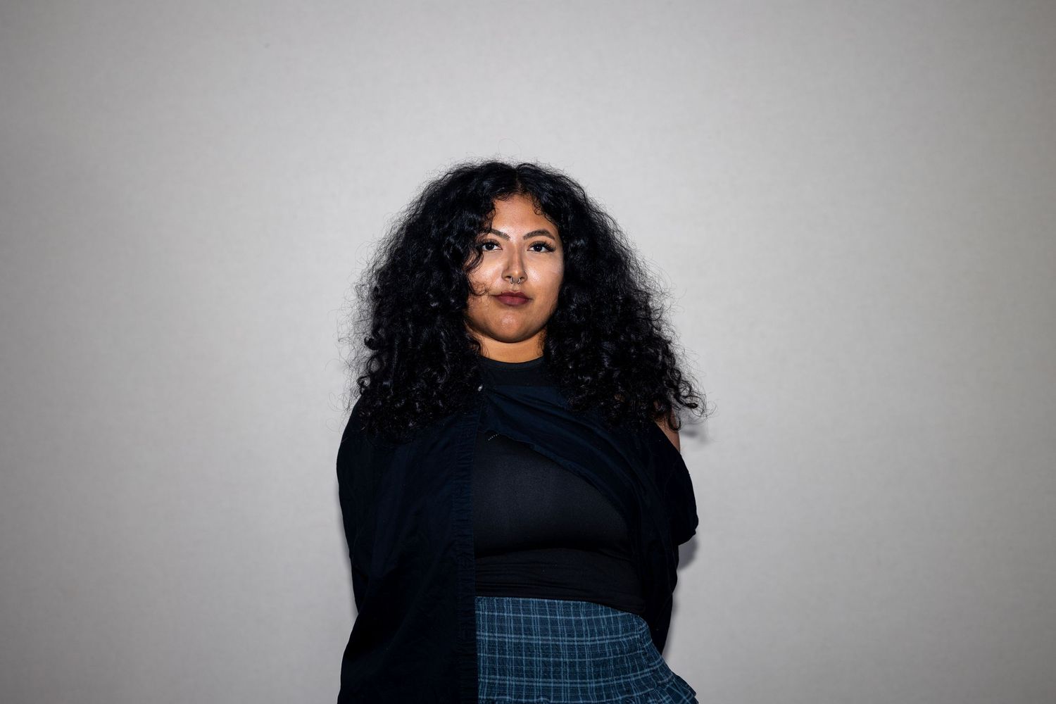 A young woman with curly black hair wearing a black top and plaid skirt poses against a gray background.
