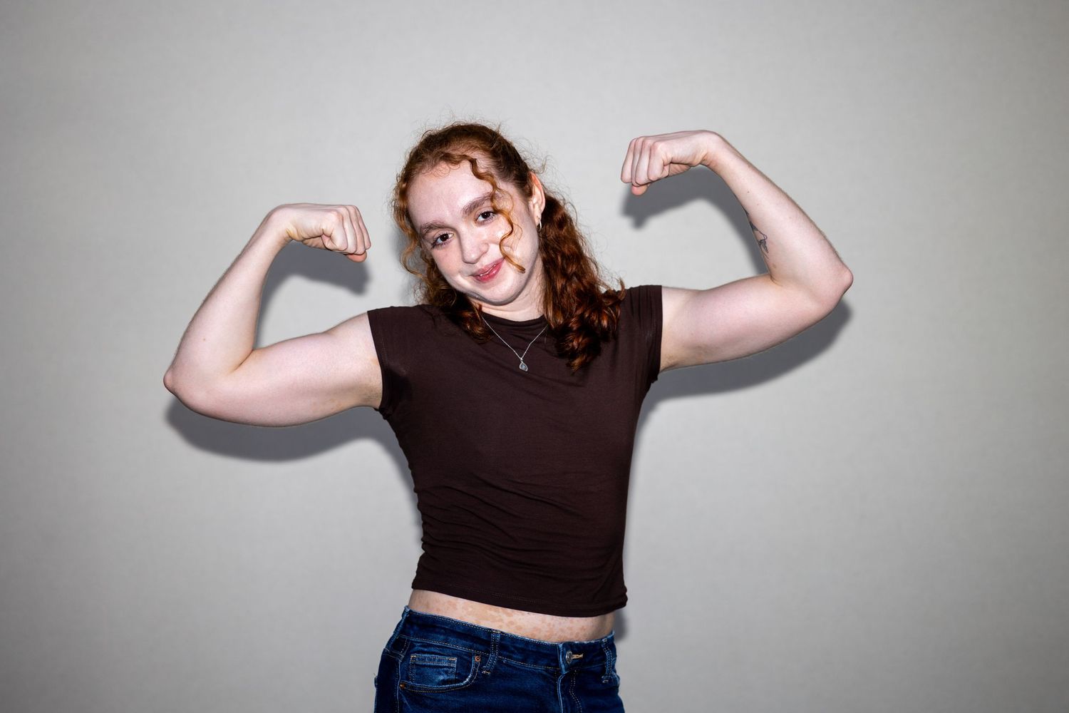 Young woman with red curly hair flexing biceps in brown crop top and jeans.