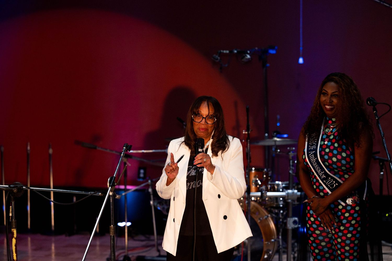 Two women on stage, one in a white blazer speaking into a microphone, the other in a polka dot dress, at a live event.