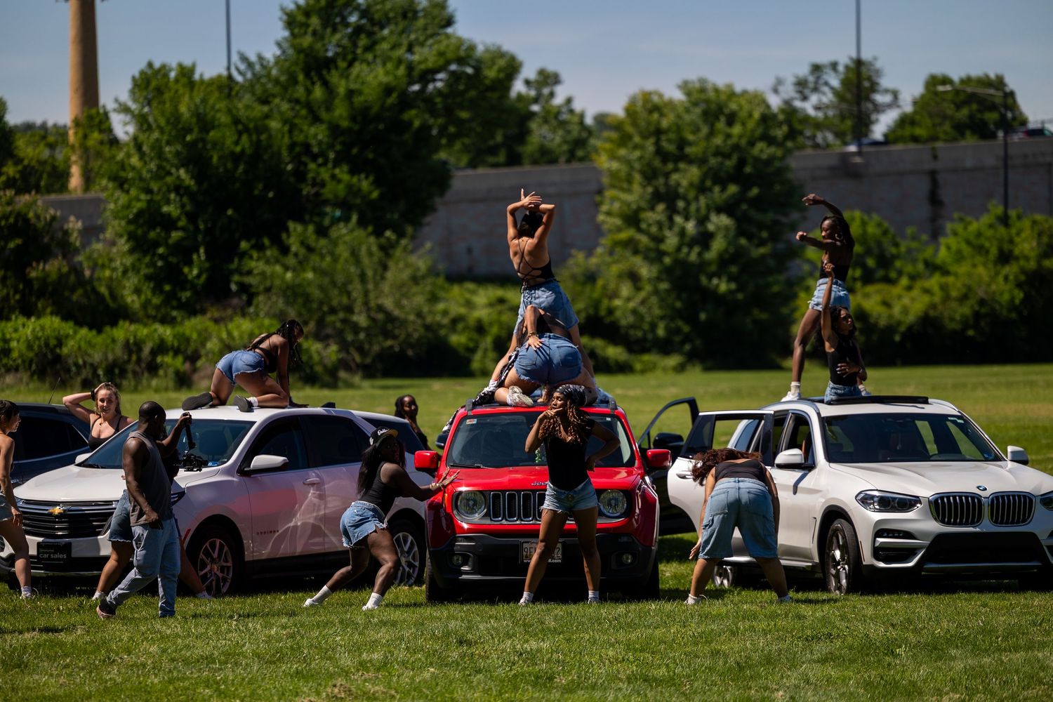 Cheerleaders perform acrobatic stunts on colorful cars parked on a green field during an outdoor event.