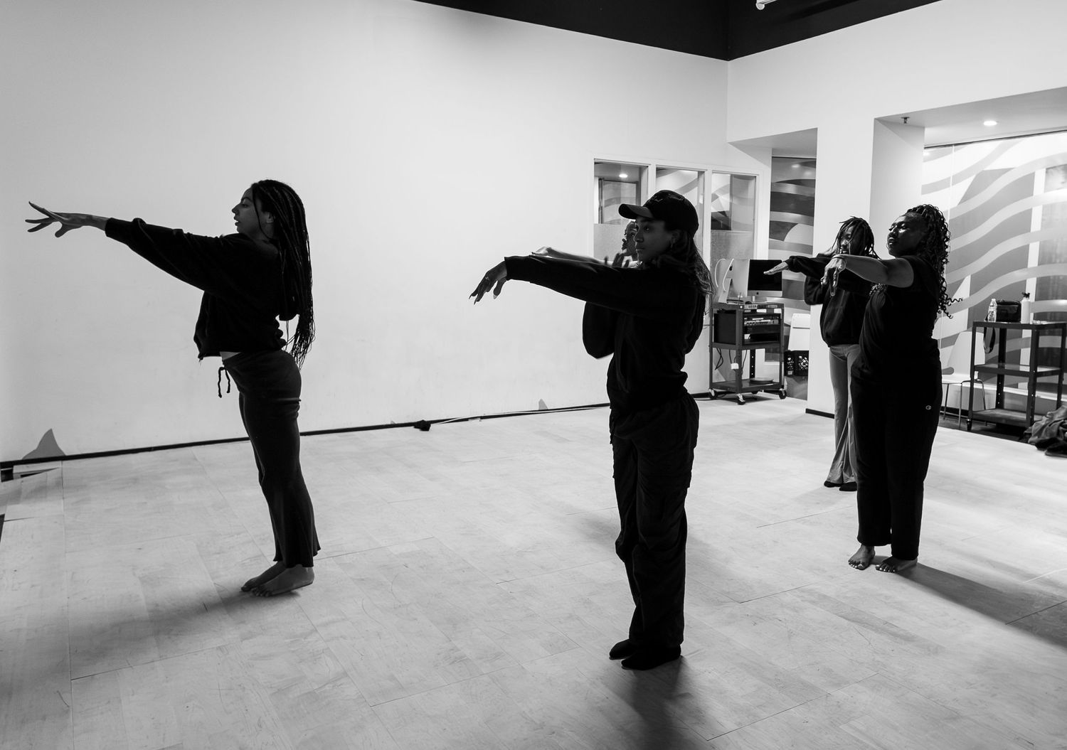 Black and white photo of four dancers rehearsing expressive choreography in a bright studio space.