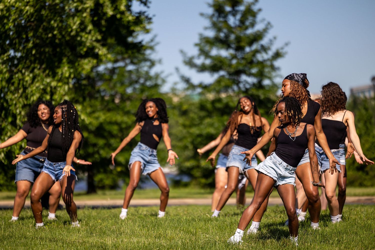 A group of young women in black tops and denim shorts dancing together on a green lawn outdoors.