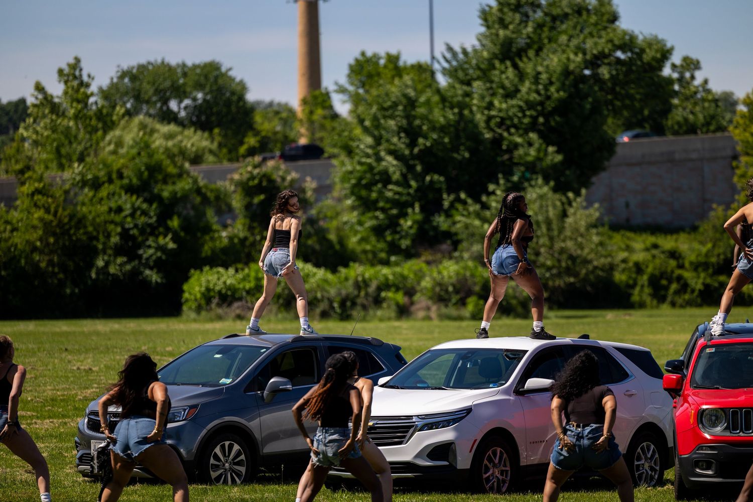 Women dancing on top of cars at an outdoor event on a green field with trees in the background.