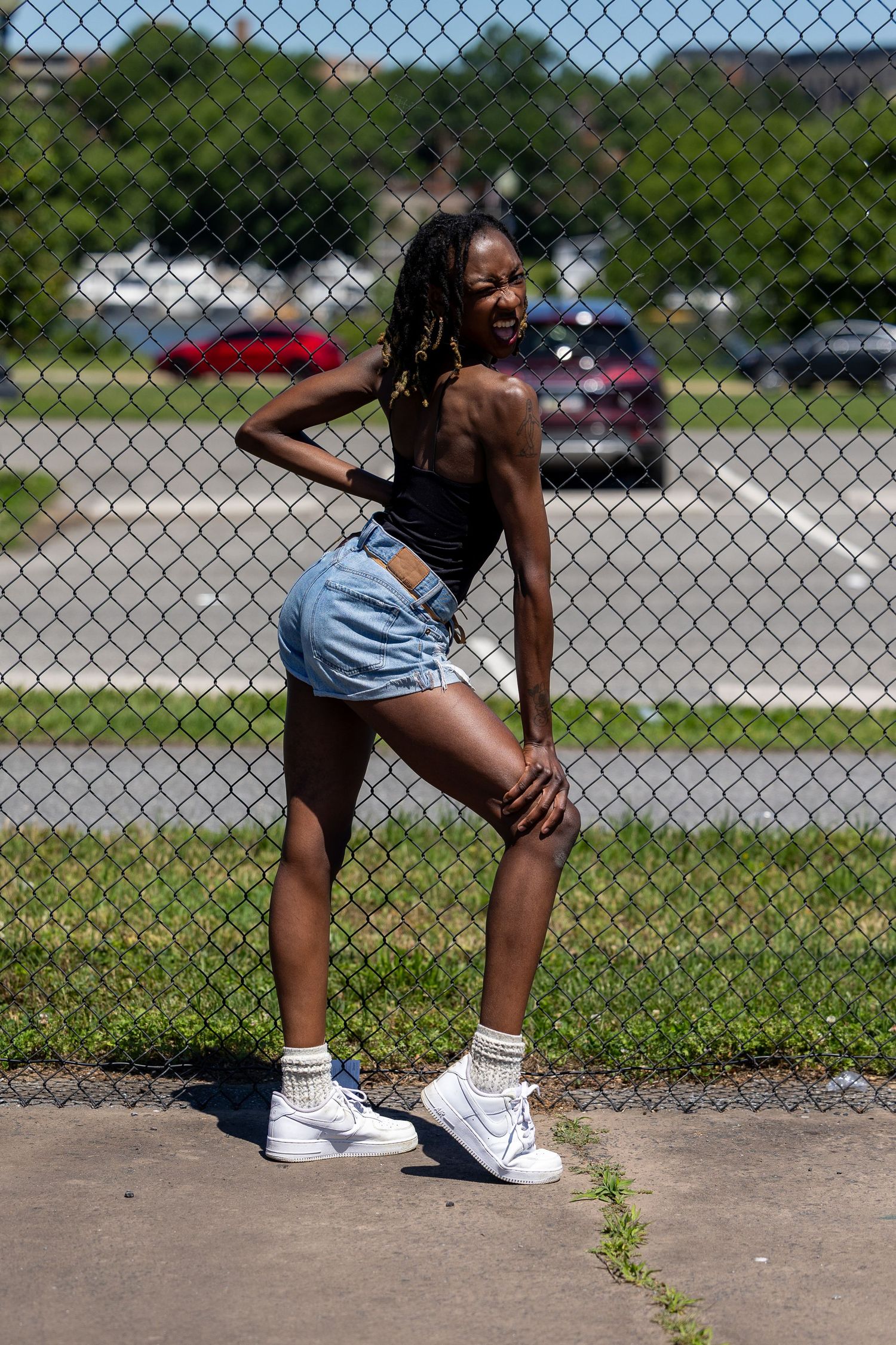 Young Black woman in denim shorts and white sneakers posing by a chain-link fence outdoors on a sunny day.