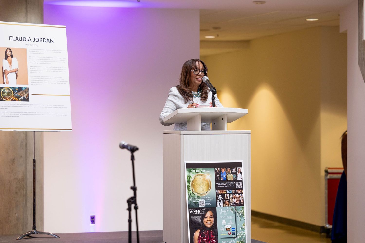 Claudia Jordan speaks at a podium during a formal event with a presentation slide displayed beside her.