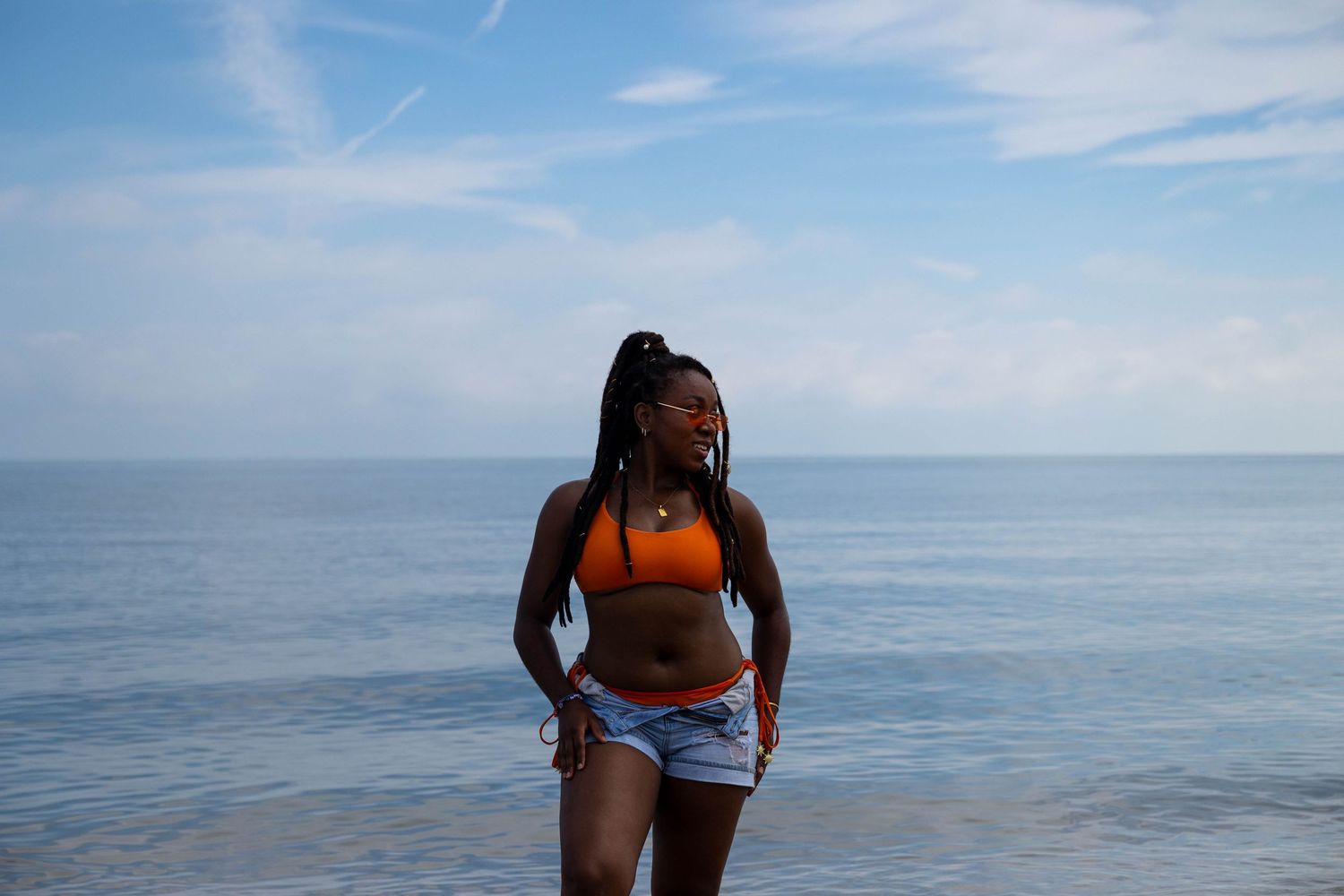 Woman in orange bikini top and denim shorts standing on beach.