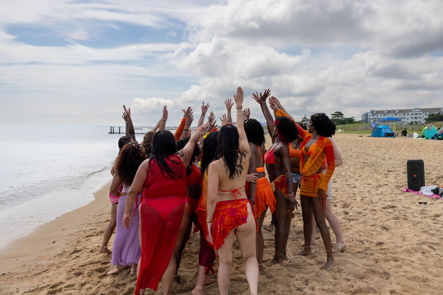 Group of women in colorful outfits raising hands on sandy beach with ocean and cloudy sky backdrop.