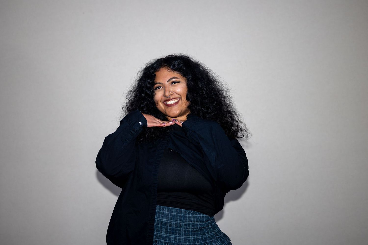 Cheerful plus-size Latina woman with curly hair wearing a navy sweater and plaid skirt, smiling warmly.