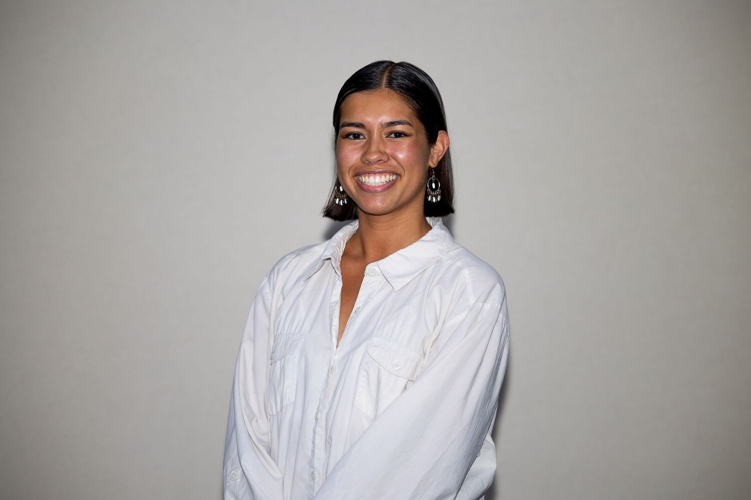 A smiling woman with sleek hair wearing a white button-down shirt and dangling earrings poses against a light wall.