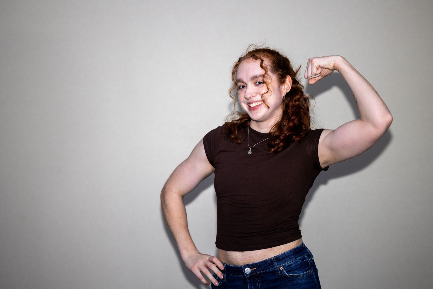 A smiling redhead teen in a brown t-shirt and jeans flexes her bicep against a gray background.