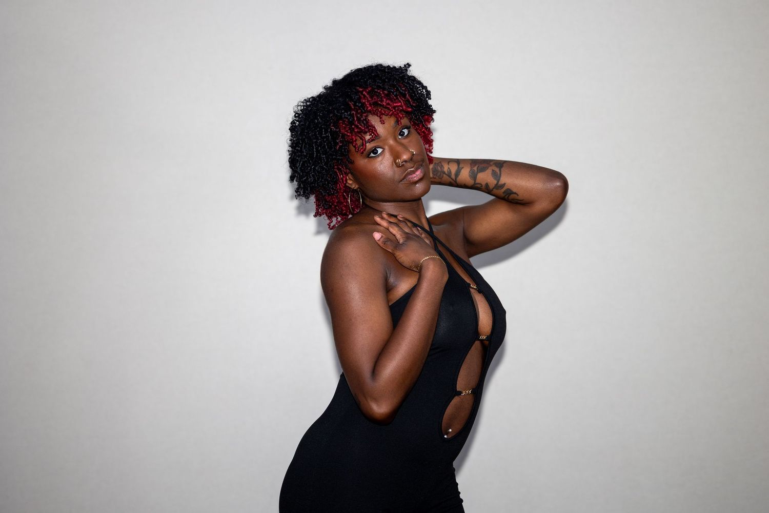 Young Black woman with red curly hair in black cutout dress posing against a light wall.