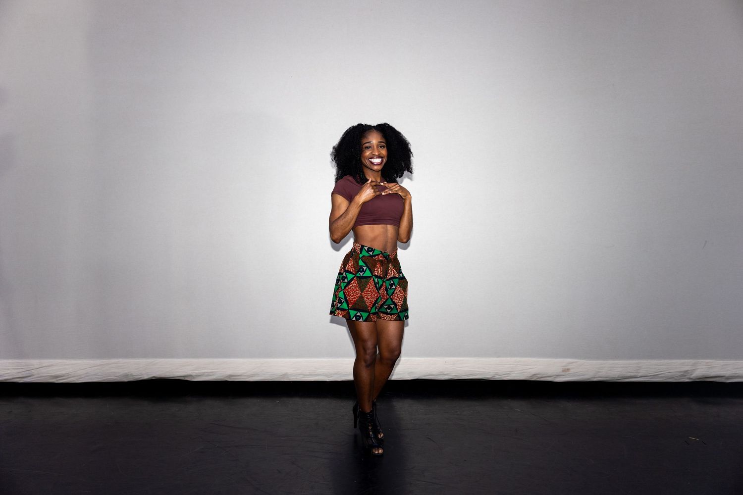 A dancer smiles in a colorful printed skirt and mauve crop top on a dark studio floor.