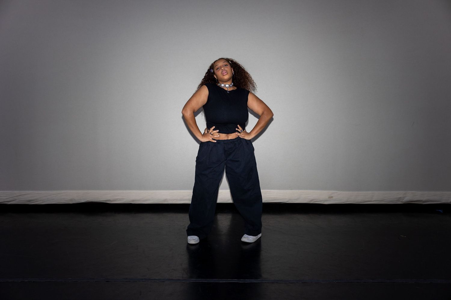 A confident dancer stands with hands on hips in a black tank and navy wide-leg pants in a studio.