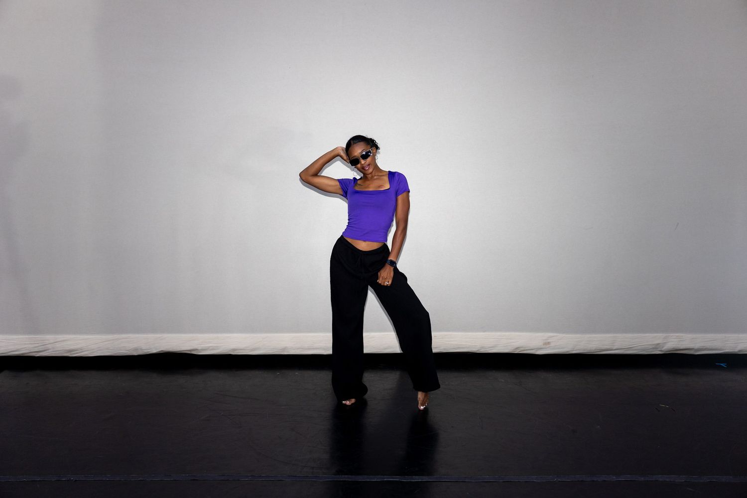 Black woman in purple crop top and black pants dances in bright studio space.