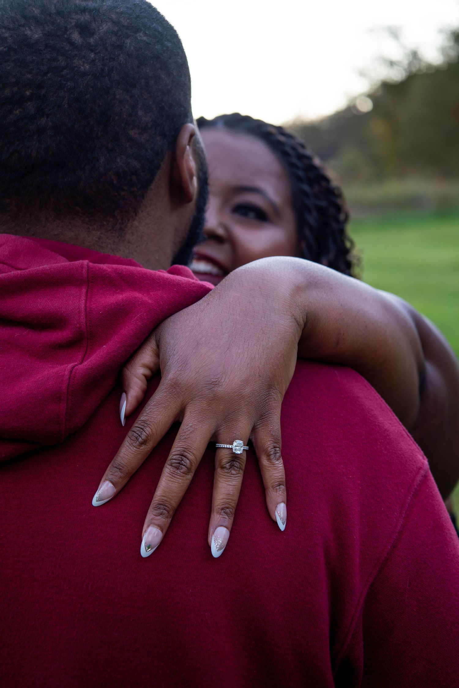 A smiling woman embraces a man outdoors, showcasing her diamond engagement ring on her hand.