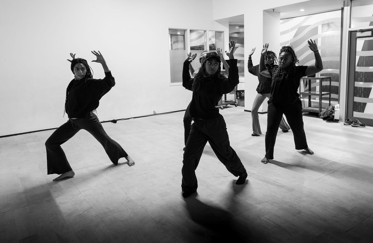 Black and white photo of four dancers in all-black outfits performing expressive choreography in a mirrored dance studio.