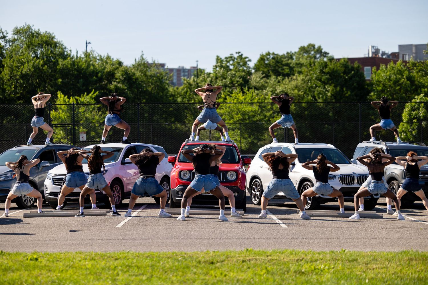 Dancers perform synchronized choreography in a parking lot surrounded by cars and green trees on a sunny day.