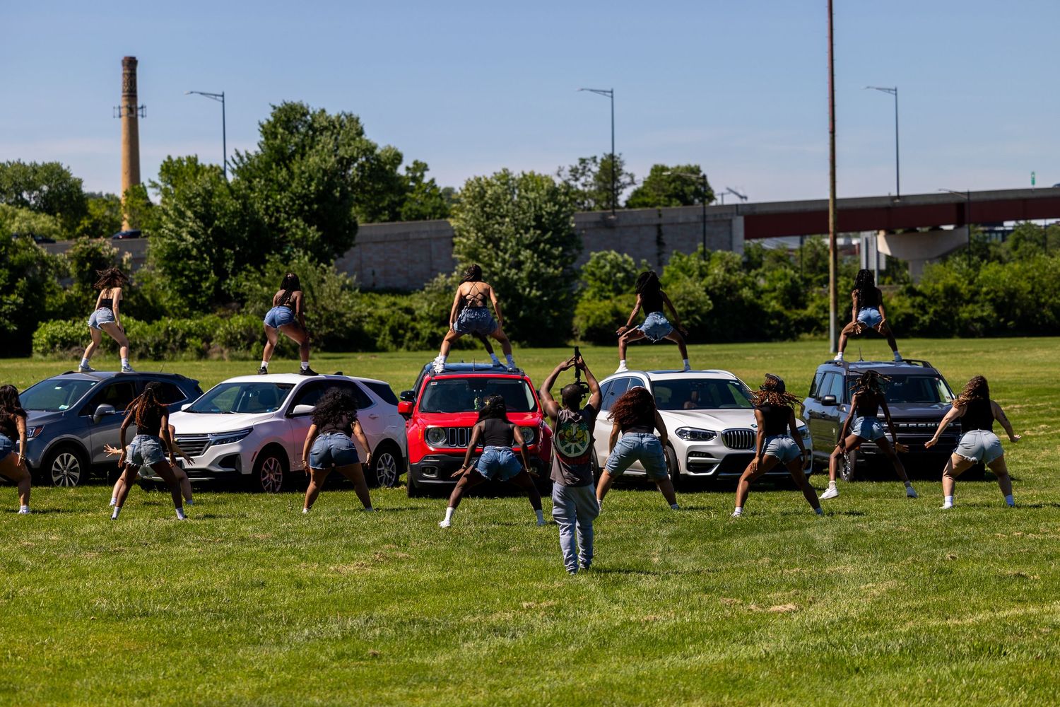 Cheerleaders performing stunts on and around parked cars on a green field during an outdoor practice session.