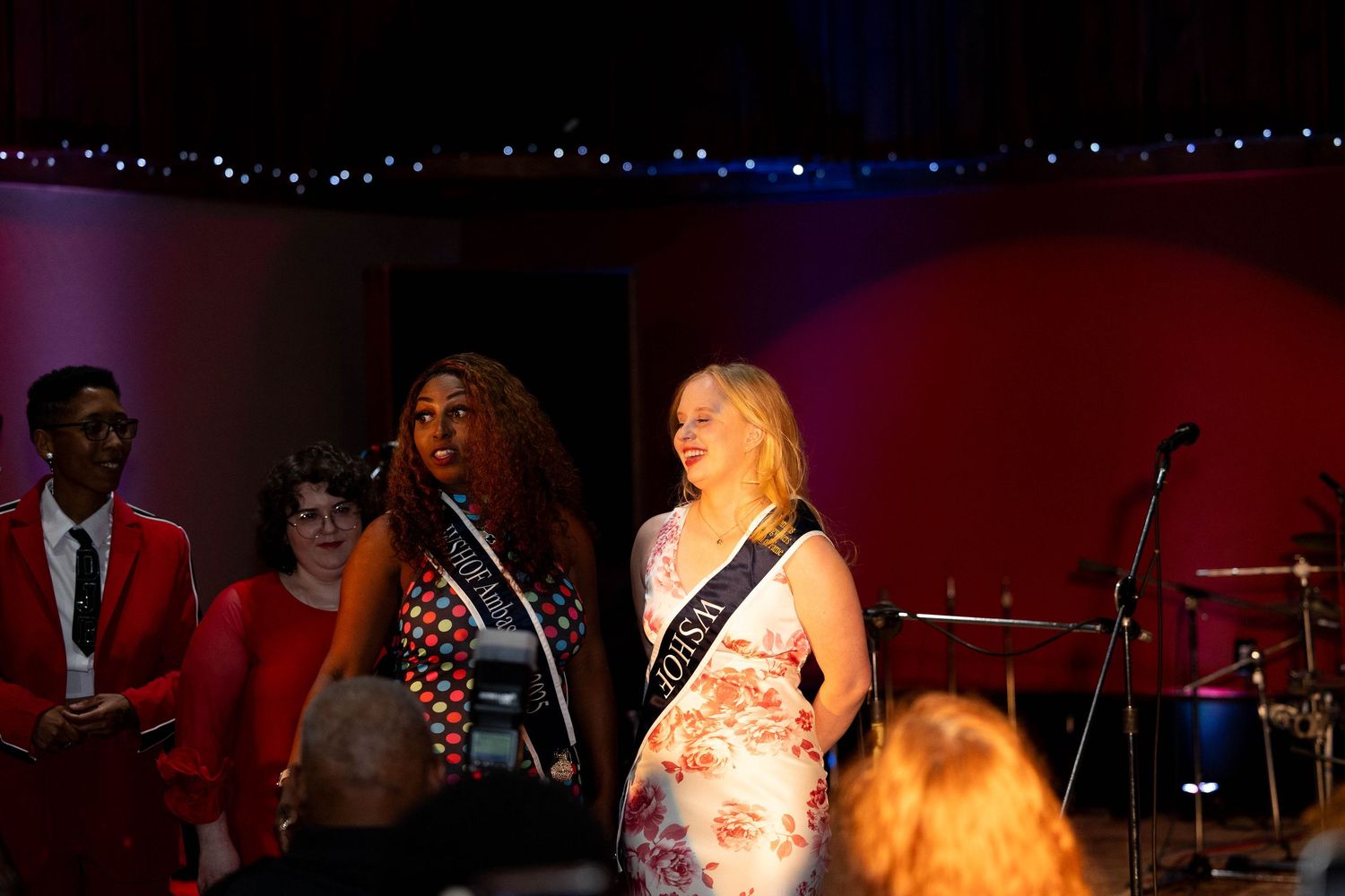 Two women wearing sashes stand on stage at a pageant or formal event with decorative lights in the background.