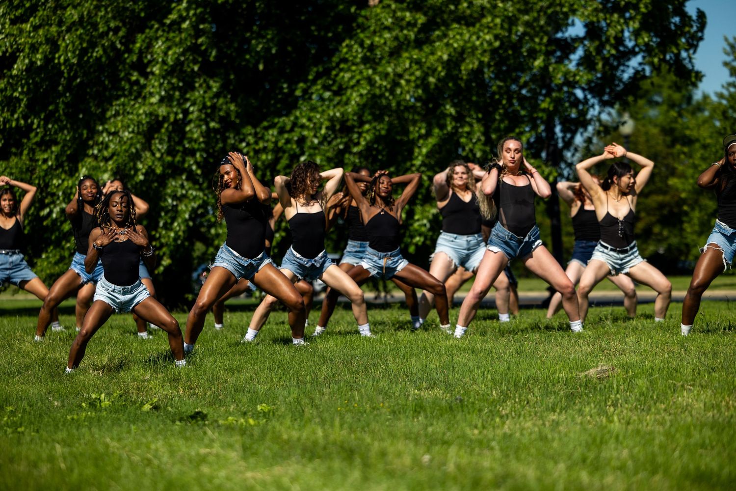 A group of young women in black tops and denim shorts perform a synchronized dance routine on a grassy park lawn.