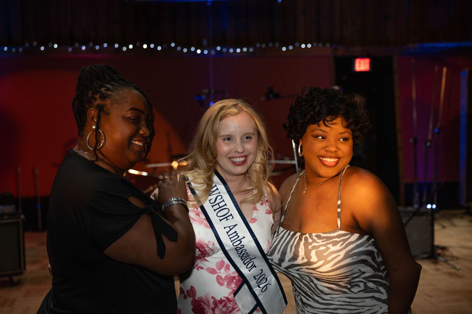 Three women posing together at a pageant event, with the middle woman wearing a Miss Ambassador sash.