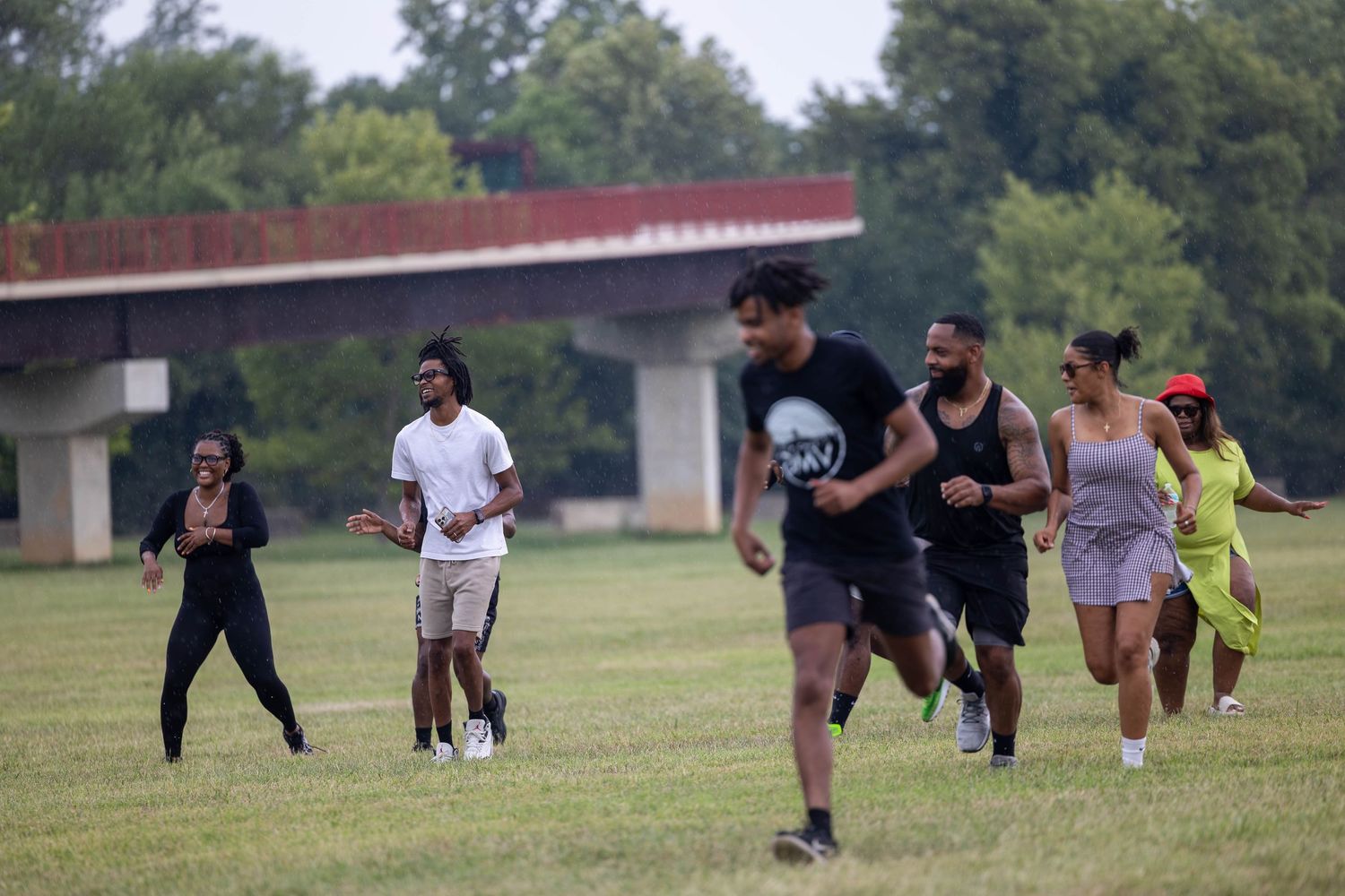 Group of people running and playing on a green grass field outdoors near a bridge on a summer day.