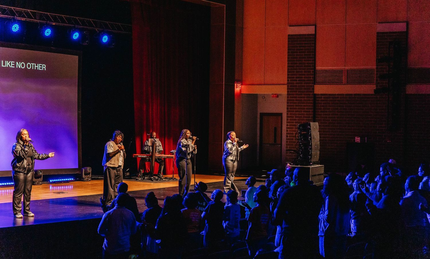A stage performance with blue lighting illuminates performers while audience members stand watching in a dark venue.