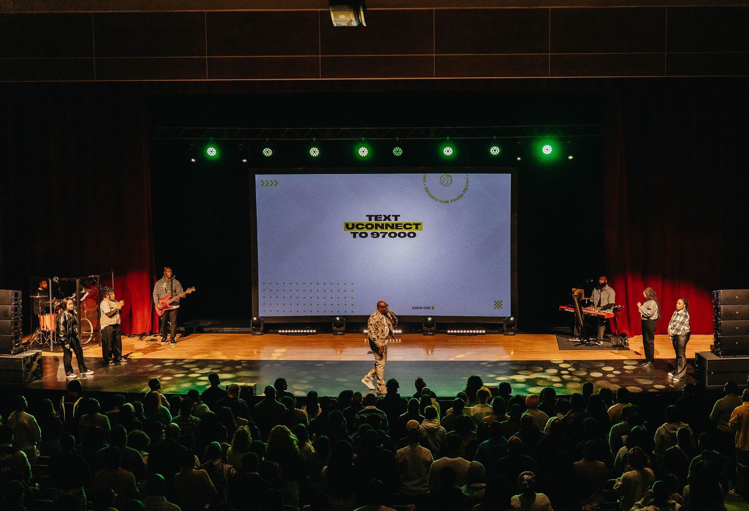 Stage performance with green spotlights and projection screen in front of seated audience in theater venue.