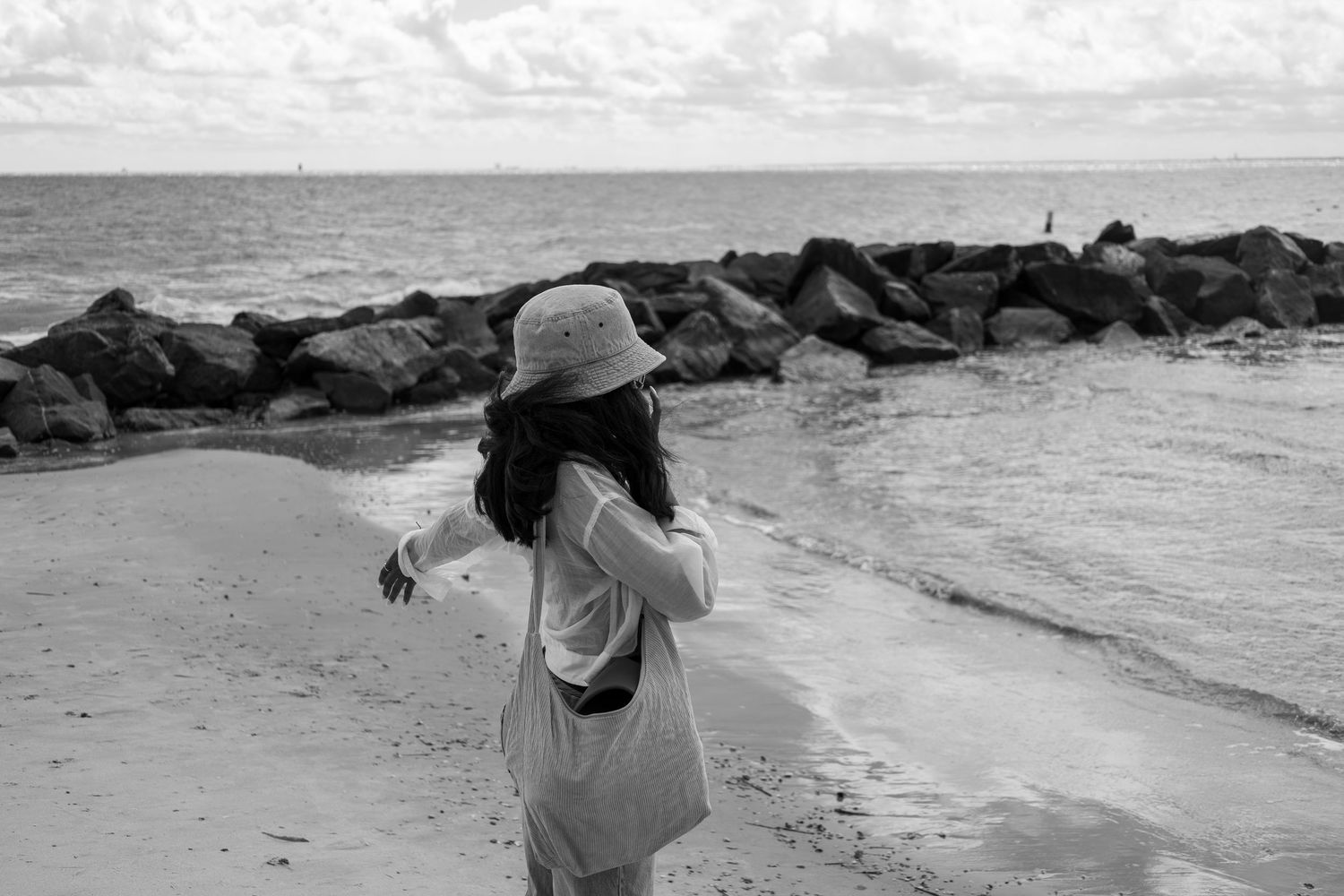 Black and white photo of someone standing on a rocky beach looking at the ocean.