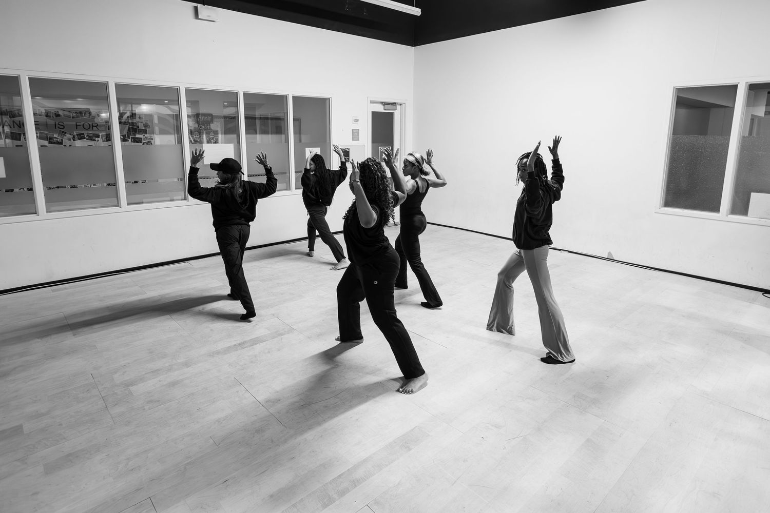 Black and white photo of a group of dancers rehearsing in a bright studio, all wearing dark clothing.