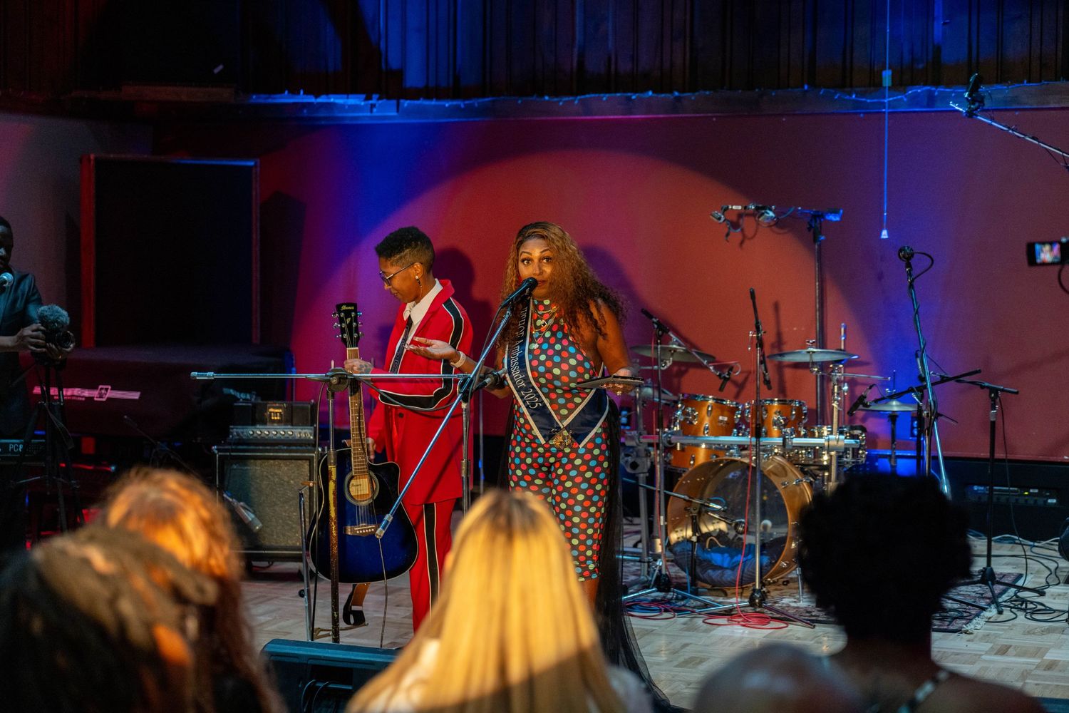 Two performers on stage, one in red jacket with guitar, other in sequined dress at live music event.