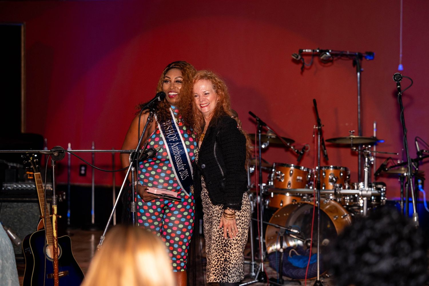 Two women pose on stage, one wearing a colorful dress with a pageant sash, the other in a black jacket and leopard skirt.