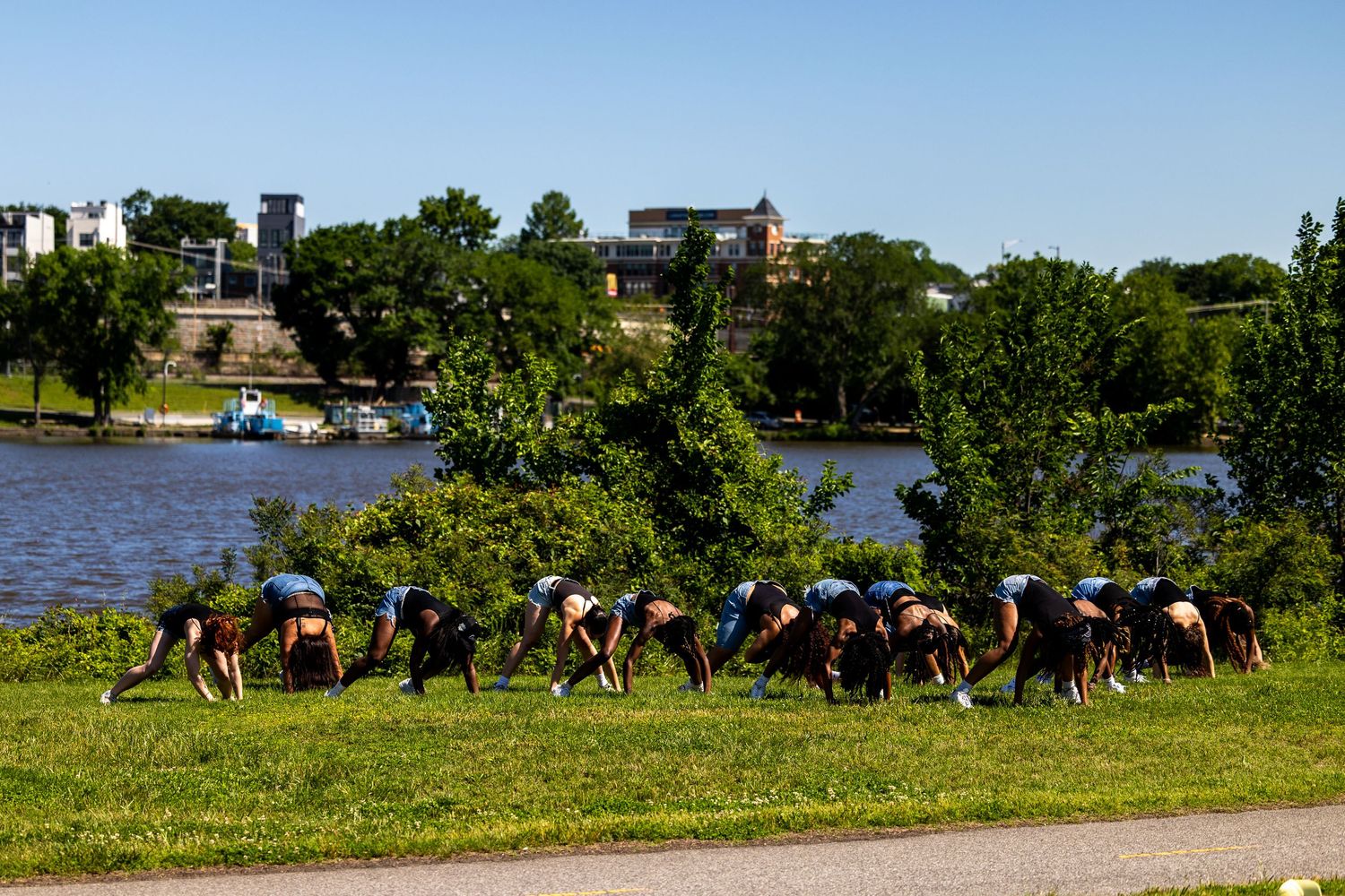Group of people doing outdoor yoga stretches on green grass beside a scenic river with trees and buildings in background.