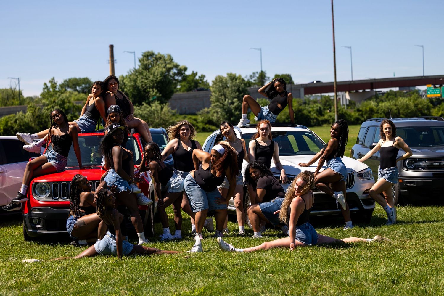 Large group of young adults posing together on cars in a sunny parking lot near a highway overpass.