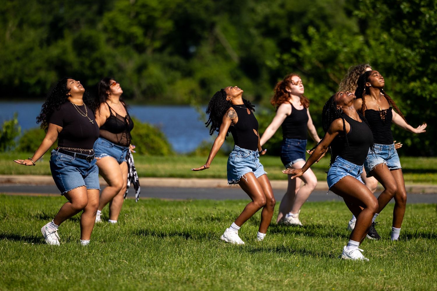 Six young women in black tops and denim shorts dance joyfully on green grass near a lake on a sunny day.