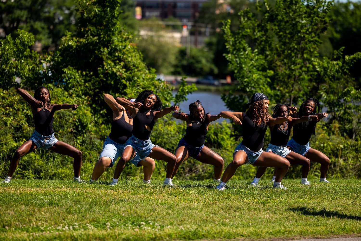 A group of young women performing a synchronized dance routine outdoors on a grassy field surrounded by trees.