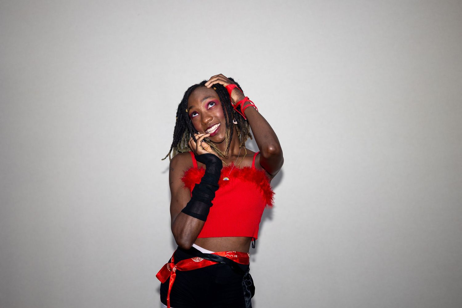 Young woman in red crop top and black fingerless gloves poses playfully against gray background.