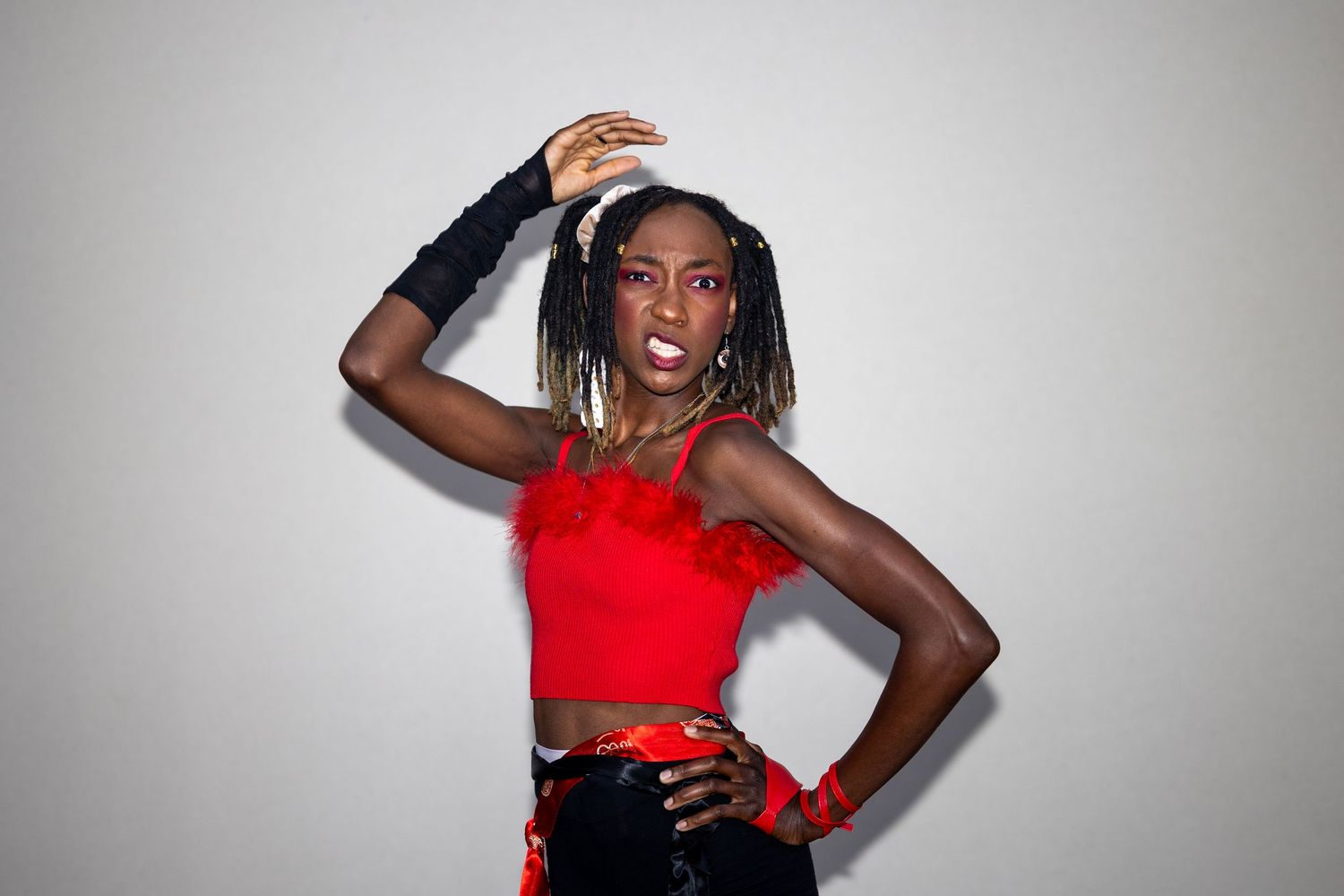Woman in red crop top and black fingerless gloves poses confidently against a light gray background.
