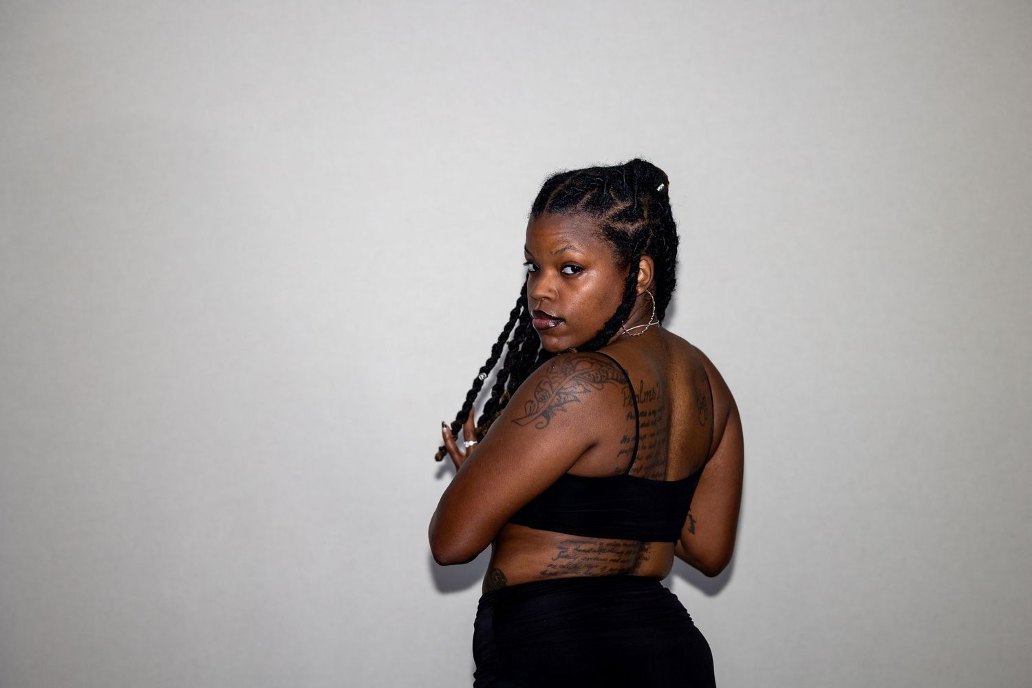 A young Black woman in a black crop top looks over her shoulder, showing tattoos on her arm.