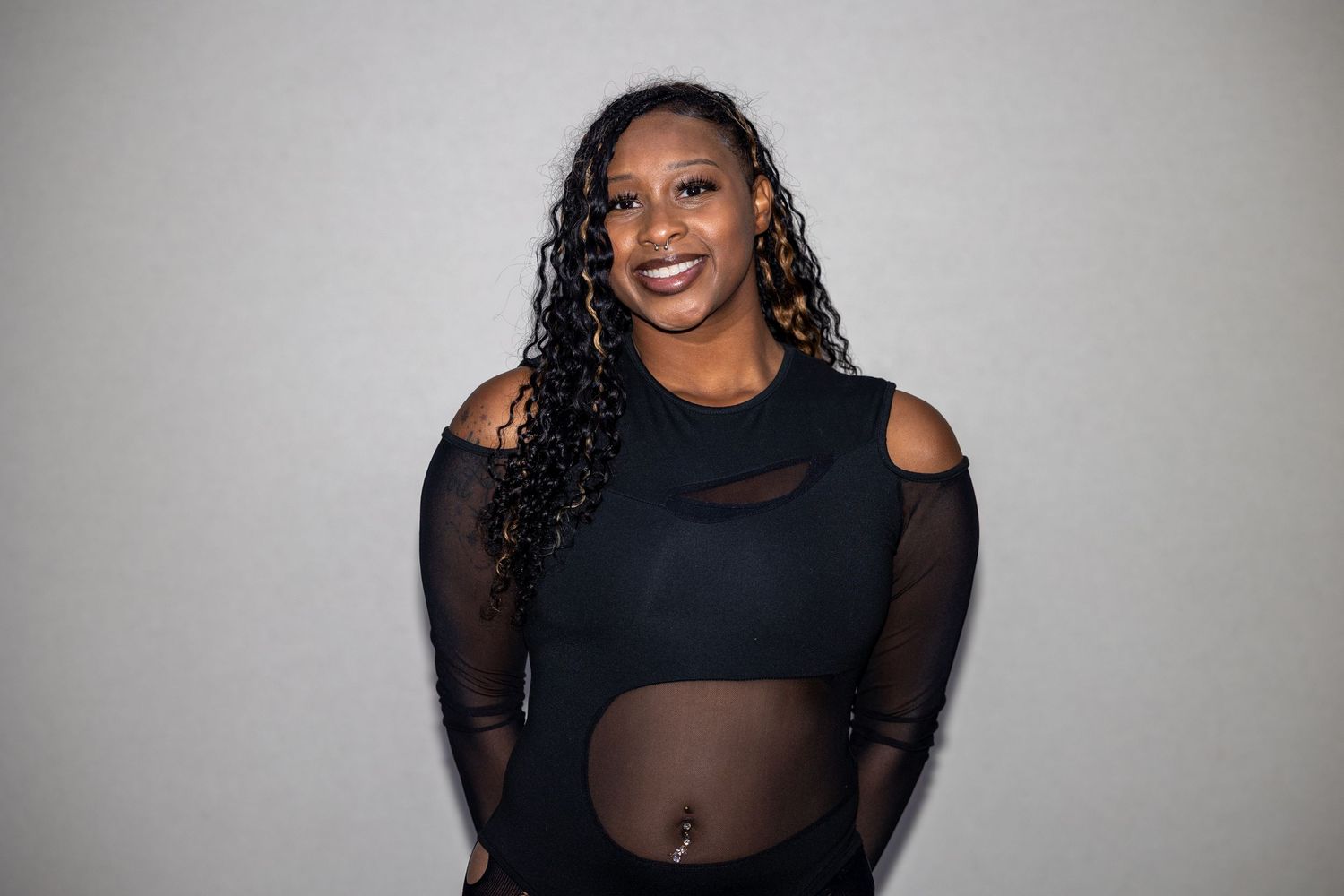 A woman in a black mesh cutout bodysuit smiles warmly while posing against a light gray background.