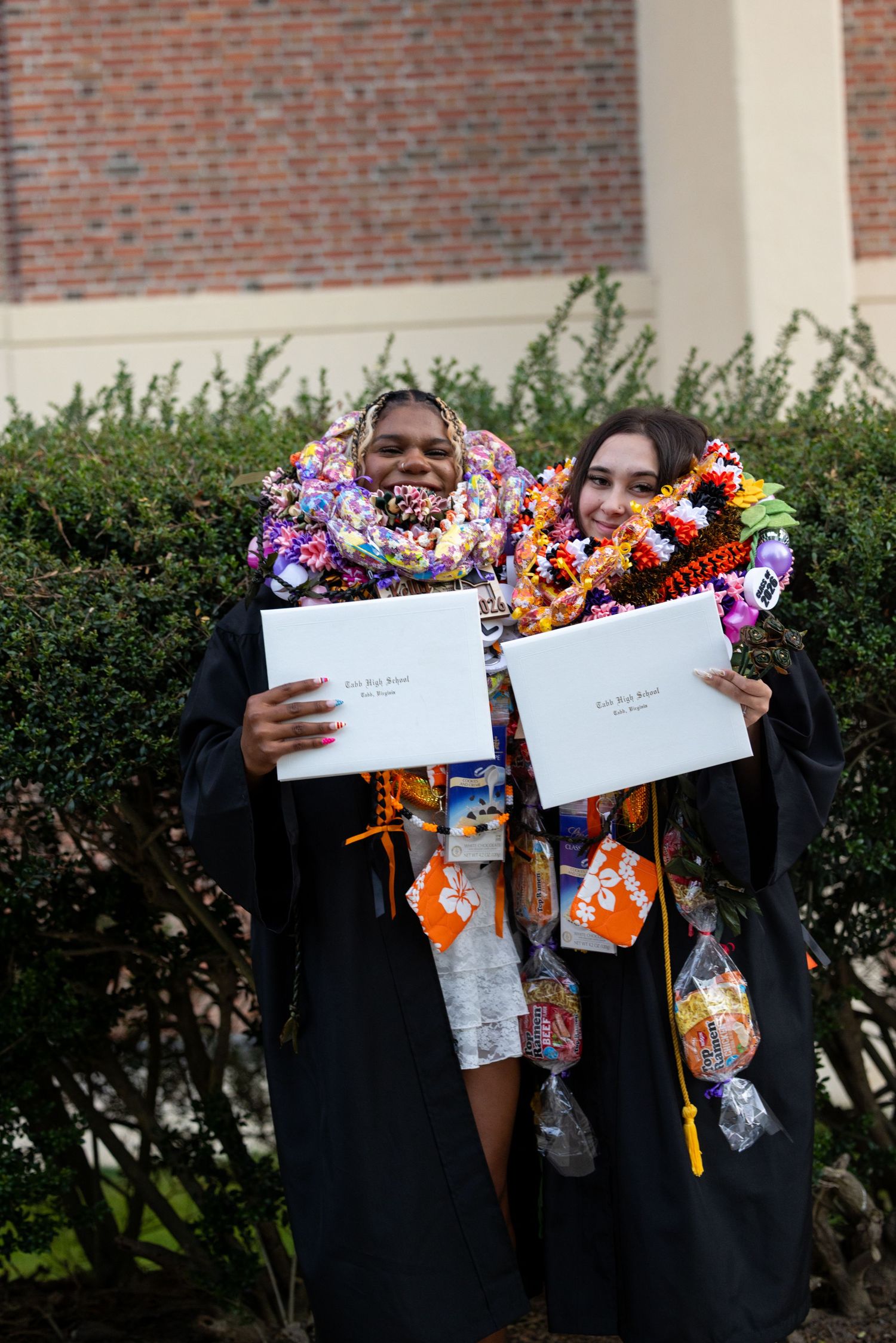 Two graduates in black gowns smile holding diplomas, adorned with colorful leis and candy necklaces outdoors.