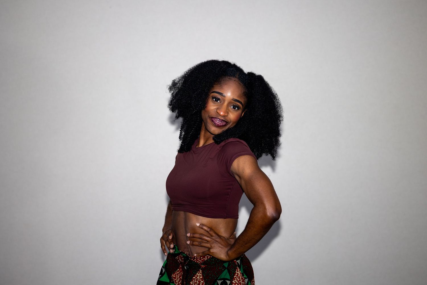 Young woman in maroon crop top and patterned skirt posing sideways with a smile.