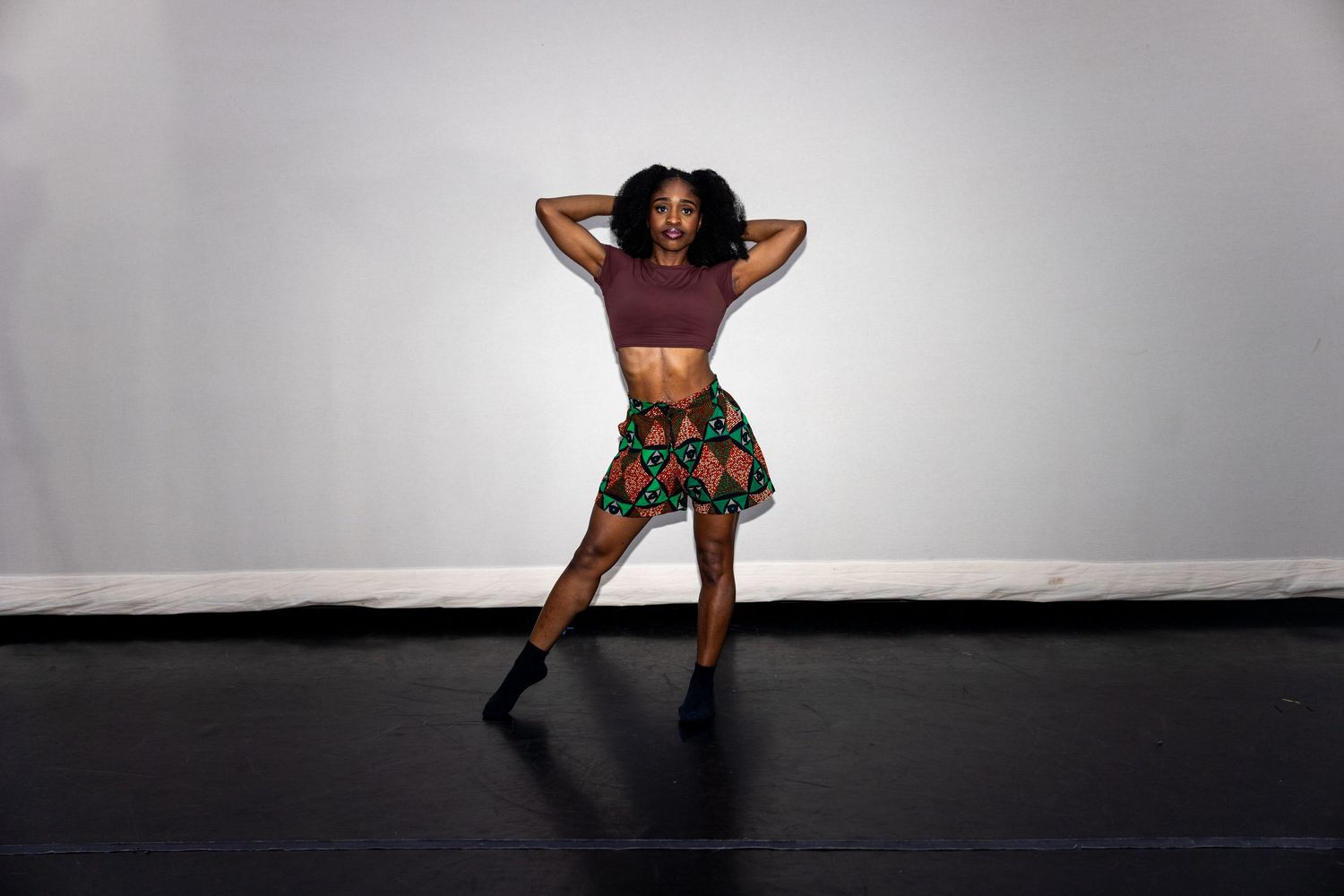 A woman in a burgundy crop top and colorful African print skirt strikes a confident dance pose on a black floor.