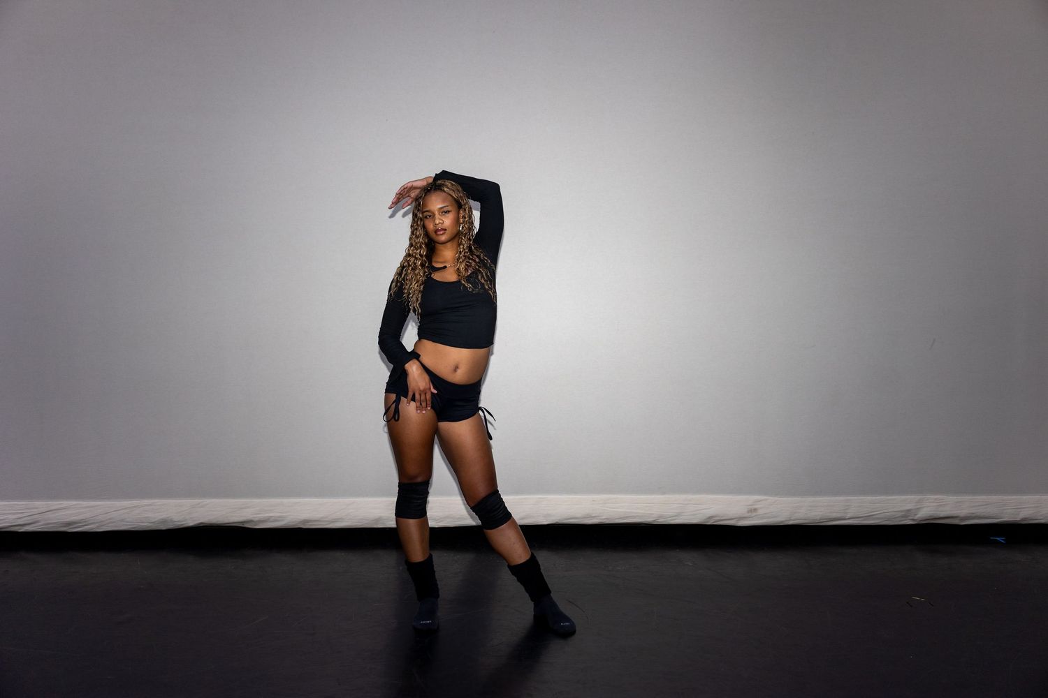 A dancer in black outfit and knee-high socks poses expressively in a professional dance studio.