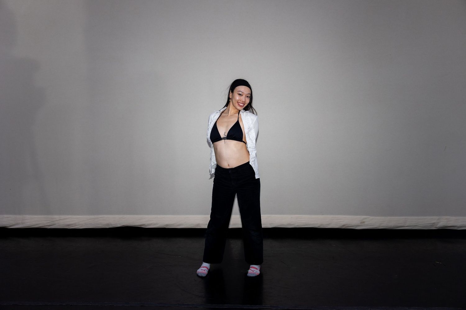Woman in white blazer and black bralette posing on dark stage floor.